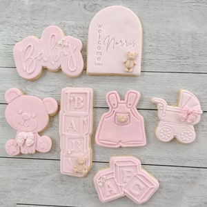 Pode incluir: Biscoitos de ch&aacute; de beb&ecirc; rosa e branco com v&aacute;rios desenhos, incluindo um urso de pel&uacute;cia, um carrinho de beb&ecirc;, um macac&atilde;o e blocos que soletram "BABY". Um biscoito tem o texto "Bem-vindo beb&ecirc; Norris".