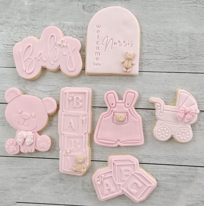 Pode incluir: Biscoitos de ch&aacute; de beb&ecirc; rosa e branco com v&aacute;rios desenhos, incluindo um urso de pel&uacute;cia, um carrinho de beb&ecirc;, um macac&atilde;o e blocos que soletram "BABY". Um biscoito tem o texto "Bem-vindo beb&ecirc; Norris".