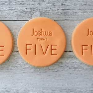 May include: Three round, peach-coloured cookies with the name "Joshua" and the words "TURNS FIVE" embossed on the surface. The cookies are arranged on a light grey wooden surface.