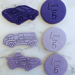 Pu&ograve; includere: Tre biscotti viola decorati con un design di auto e il testo "Louis compie 5 anni".