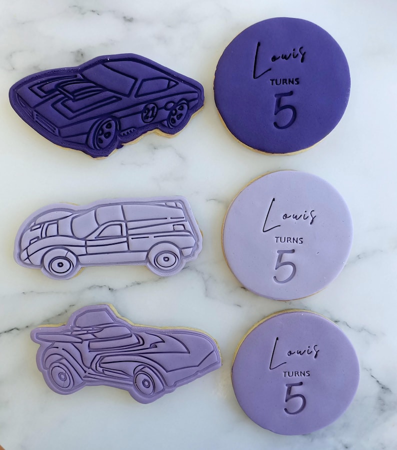 Pu&ograve; includere: Tre biscotti viola decorati con un design di auto e il testo "Louis compie 5 anni".