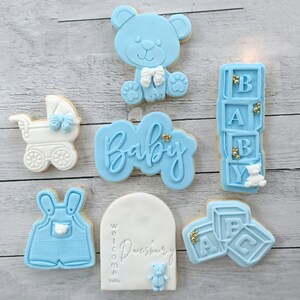 Pode incluir: Um conjunto de seis bolachas decoradas em azul e branco. As bolachas t&ecirc;m o formato de um urso de peluche, um carrinho de beb&eacute;, um conjunto de blocos, um macac&atilde;o, uma placa de boas-vindas e a palavra "Beb&eacute;".