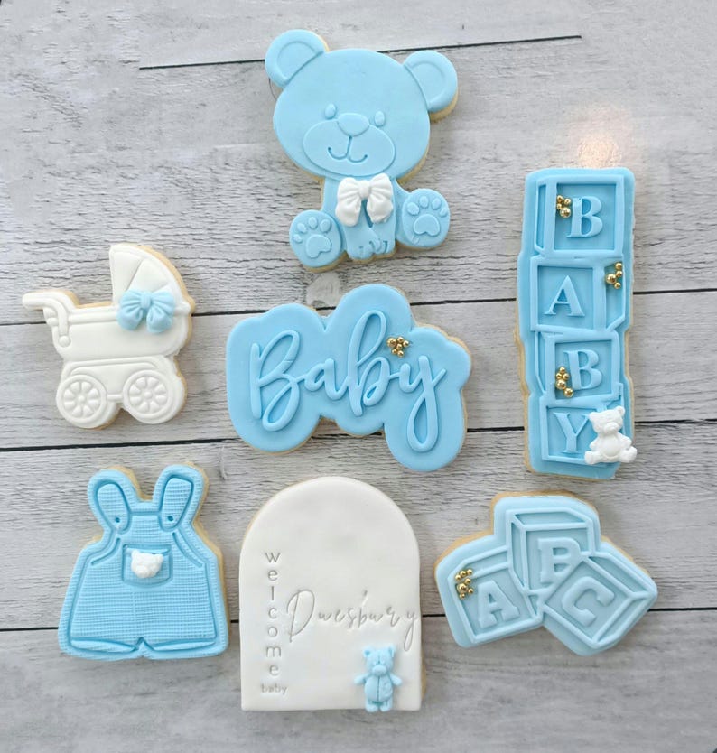 Pode incluir: Um conjunto de seis bolachas decoradas em azul e branco. As bolachas t&ecirc;m o formato de um urso de peluche, um carrinho de beb&eacute;, um conjunto de blocos, um macac&atilde;o, uma placa de boas-vindas e a palavra "Beb&eacute;".