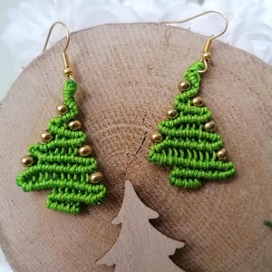 Könnte beinhalten: Ein Paar grüne Weihnachtsbaum-Ohrringe mit goldenen Perlen. Die Ohrringe hängen an einer Holzoberfläche mit einem geschnitzten Weihnachtsbaum aus Holz.