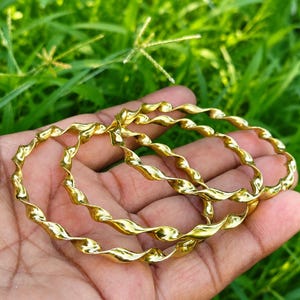 Puede incluir: Tres brazaletes dorados con un diseño continuo de espiral retorcida. Cada pulsera circular tiene una superficie pulida y reflectante que capta la luz, creando una serie de curvas repetitivas a lo largo de toda la longitud de las bandas metálicas.