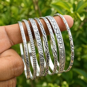 Puede incluir: Un conjunto de siete brazaletes de metal plateado con varios patrones en relieve, incluyendo motivos florales, texturas trenzadas, líneas onduladas y formas de aves repetidas. Cada brazalete tiene un diseño distinto con detalles oxidados.