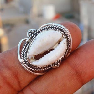 May include: A silver ring featuring a white cowrie shell set in the centre. The ring is adorned with a silver rope design around the edge.
