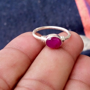 May include: A silver ring featuring a single, round, faceted, deep pinkish-red gemstone set in a simple metal band. The setting includes small, decorative horizontal ridges on either side of the stone, which is polished and reflective.