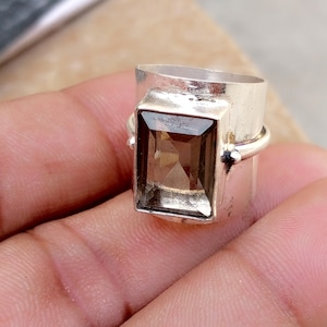 May include: A wide silver band ring featuring a large, rectangular, faceted smoky quartz gemstone set in a polished metal bezel. The ring has a smooth, metallic finish with a prominent, clear, brown-toned stone centered on the front surface.
