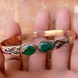 May include: A copper cuff bracelet featuring two teardrop-shaped green gemstones at the open ends. The metal band has a smooth finish, while the settings for the stones are decorated with intricate twisted wire and small metal bead accents.