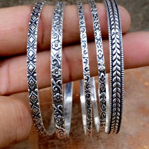 Pode incluir: Cinco pulseiras de metal prateado com padrões em relevo distintos. Os designs incluem formas geométricas de diamante, motivos florais, volutas intrincadas e um padrão repetitivo de folhas, tudo com um acabamento oxidado para realçar os detalhes.