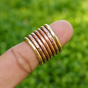 May include: A stack of seven thin metal rings worn on a finger. The set includes alternating bands of polished gold-tone metal and textured, hammered copper-tone metal, creating a layered appearance with varying metallic finishes and surfaces.