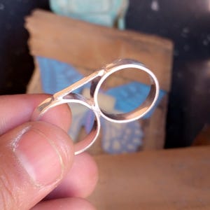 May include: A silver double ring with two connected circles. The ring is worn on a finger.