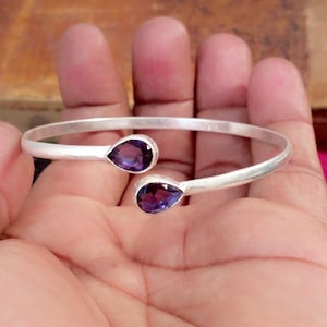 Peut inclure: Un bracelet manchette argenté avec deux pierres précieuses en forme de goutte d'eau, de couleur violet foncé. Le bracelet a un design simple et élégant, parfait pour un usage quotidien. Les pierres sont serties à chaque extrémité du bracelet ouvert.