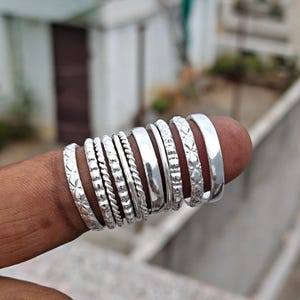 May include: A collection of nine silver-toned rings arranged in a row. The set includes various designs such as smooth bands, twisted rope textures, beaded patterns, and intricate geometric engravings, all displayed together on a finger.