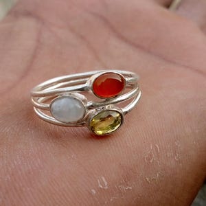 May include: A stackable ring set with three silver bands, each featuring an oval gemstone. The gemstones are a vibrant orange, a pale white, and a yellow-green color. The rings are set on a hand.