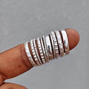 May include: A set of ten silver-toned rings featuring various textures and patterns, including twisted rope designs, smooth polished bands, and intricate geometric engravings, all arranged in a row on a finger for display and comparison.