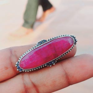 以下が含まれることがあります： ビーズのベゼルにセットされた、大きな楕円形のピンクの宝石が付いたシルバーリングです。