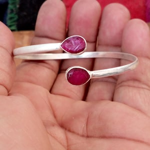 May include: A silver cuff bracelet with two teardrop-shaped, deep red gemstones. The bracelet has an open design, with the gemstones set at each end of the band. The gemstones are a vibrant, saturated colour, contrasting with the polished metal.