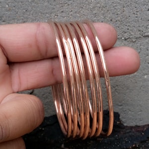 Op de afbeelding: Een stapel koperkleurige armbanden. De armbanden zijn rond en lijken van dun metaal te zijn gemaakt. De armbanden zijn op elkaar gestapeld en worden door een hand vastgehouden.