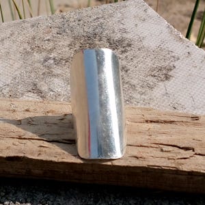 May include: A silver-colored ring with a rectangular design. The ring is made of a shiny metal and has a smooth surface. The ring is displayed on a piece of wood.