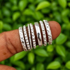 Puede incluir: Un conjunto de siete anillos de color plateado con diversas texturas, incluyendo bandas con patrón de diamantes, un diseño de cuerda trenzada, bandas con cuentas y una banda lisa y pulida. Los anillos están dispuestos uno al lado del otro en un dedo, mostrando acabados metálicos distintos y detalles de superficie intrincados.