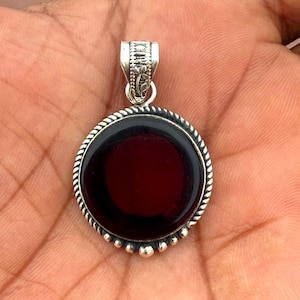 May include: A silver pendant featuring a circular, deep red gemstone. The stone is set within a silver bezel, adorned with a rope-style edge and tiny silver beads. The bail is decorated with floral designs.
