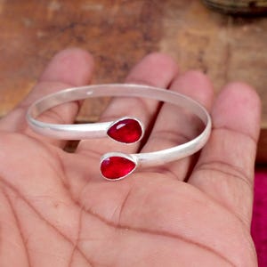 Peut inclure: Un bracelet ouvert de couleur argentée doté de deux pierres rouges en forme de goutte à chaque extrémité. La bande présente une finition mate et lisse, et les deux pierres rouges sont facettées, créant une surface réfléchissante à l'ouverture.