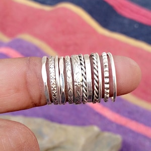 Puede incluir: Una colección de anillos de plata con varios diseños, incluyendo bandas texturizadas, trenzadas y estampadas. Los anillos se muestran en un dedo, mostrando sus diferentes estilos y acabados. Los anillos varían en ancho.