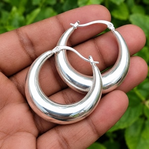 May include: A pair of polished silver hoop earrings with a thick, rounded, crescent-shaped design. The earrings feature a smooth, reflective metallic surface and a hinged post closure mechanism, showcasing a simple and classic circular silhouette.
