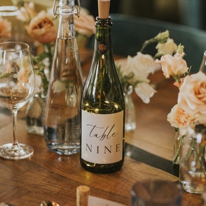 May include: A close-up of a table setting with a bottle of wine with a label that says "table nine". There are also two glasses, a water bottle, and flowers on the table.