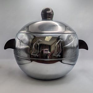 May include: A spherical chrome ice bucket featuring a polished silver finish with etched penguin motifs around the center. The container includes two dark brown side handles and a matching dark brown spherical knob on the removable lid.