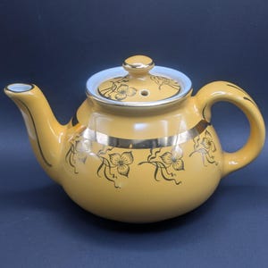 May include: Mustard yellow ceramic teapot with a gold band and floral design. The lid is white with a gold knob. The teapot has a curved handle and spout, perfect for serving tea.