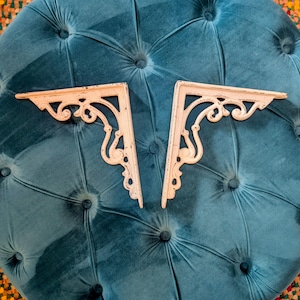 May include: Two off-white, decorative shelf brackets with ornate scrollwork. The brackets are made of metal and have a distressed finish, suggesting a vintage or antique style. They are positioned on a tufted teal surface.