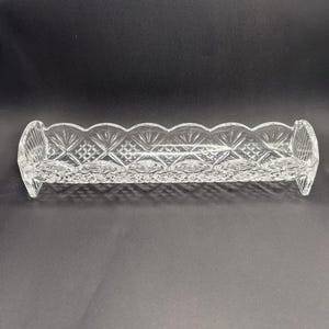 Op de afbeelding: Een heldere, rechthoekige glazen schaal met een geschulpte rand en een ruitpatroon. De schaal heeft gebogen uiteinden en is ontworpen voor kleine items. Het transparante glas laat licht door, tegen een donkere achtergrond.