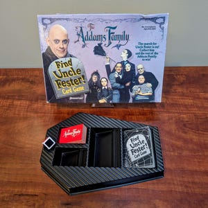 May include: The Addams Family Find Uncle Fester! card game box featuring the cast and a black plastic game tray with card slots, a deck of red-backed cards, a deck of game cards with spiderweb graphics, and a small black and white square token.