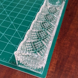 Op de afbeelding: Een heldere, rechthoekige kristallen vaas met een gedetailleerd, geometrisch patroon. De vaas heeft een reeks verbonden, cilindrische openingen en ligt op een groene snijmat. De vaas is ontworpen om meerdere stelen te bevatten.