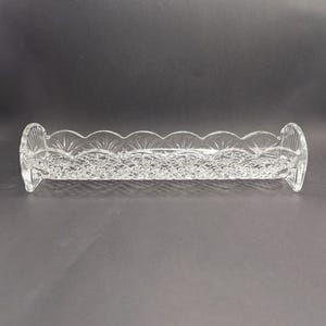 Op de afbeelding: Een heldere, rechthoekige kristallen schaal met een geschulpte rand en een ingewikkeld bloemmotief. De schaal heeft afgeronde uiteinden en is ontworpen om kleine items in te bewaren. Het kristal reflecteert licht, waardoor een sprankelend effect ontstaat.
