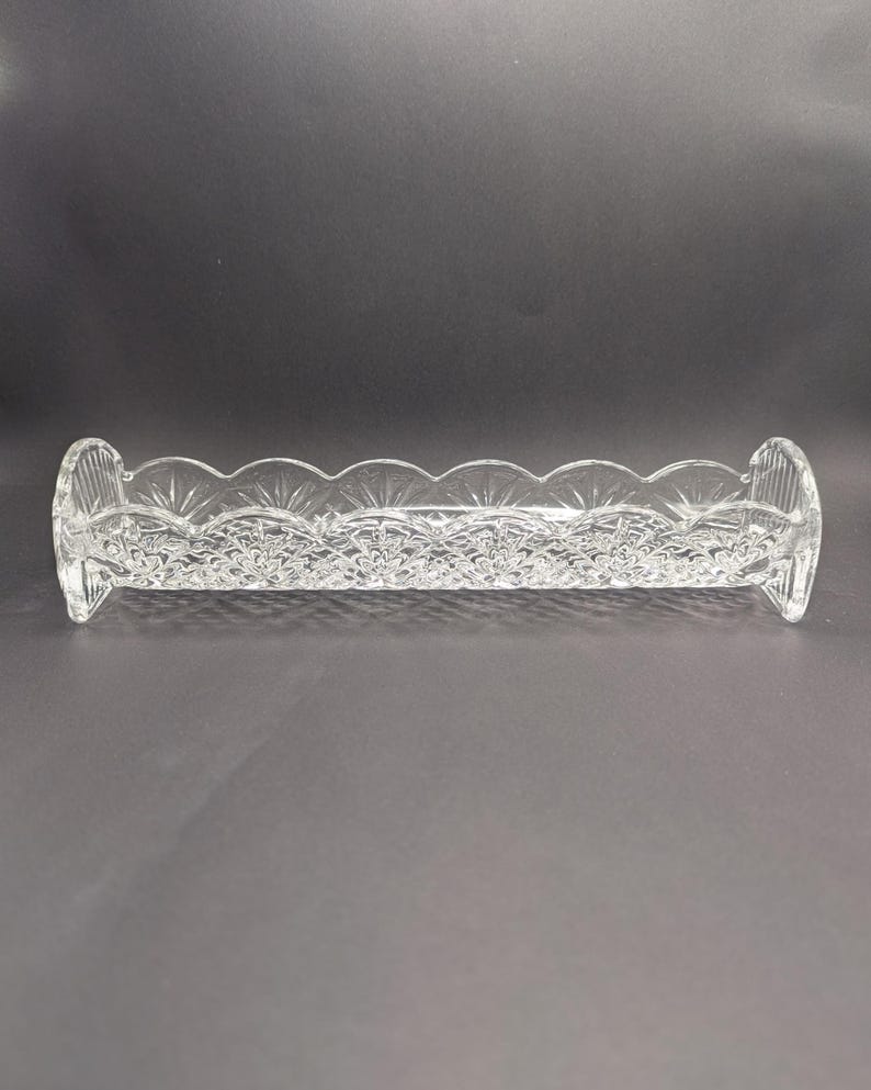Op de afbeelding: Een heldere, rechthoekige kristallen schaal met een geschulpte rand en een ingewikkeld bloemmotief. De schaal heeft afgeronde uiteinden en is ontworpen om kleine items in te bewaren. Het kristal reflecteert licht, waardoor een sprankelend effect ontstaat.