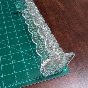 Op de afbeelding: Een helder, decoratief glazen object met een geschulpte rand. Het object heeft een reeks zich herhalende, waaiervormige elementen en ligt op een groene snijmat. Het object ligt op een donkerbruin oppervlak.