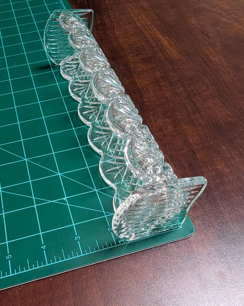 Op de afbeelding: Een helder, decoratief glazen object met een geschulpte rand. Het object heeft een reeks zich herhalende, waaiervormige elementen en ligt op een groene snijmat. Het object ligt op een donkerbruin oppervlak.