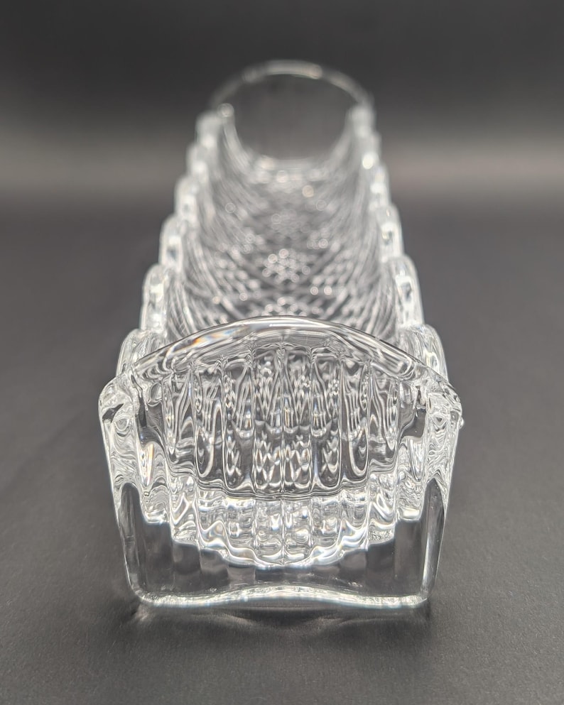 Op de afbeelding: Een heldere, rechthoekige kristallen vaas met een gestructureerd, geometrisch patroon. De vaas heeft een gebogen, open bovenkant en een reeks ribbels langs de zijkanten. Het kristal reflecteert licht en cre&euml;ert een sprankelend effect.