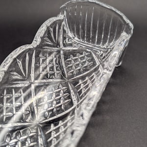 Op de afbeelding: Een heldere, decoratieve glazen schaal met een ruit- en kruispatroon. De schaal heeft een gebogen, langwerpige vorm met een geschulpte rand en een getextureerd oppervlak. De schaal is ontworpen om kleine items in te bewaren.