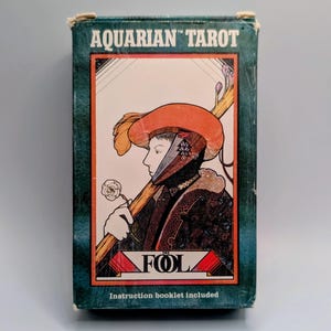 May include: A rectangular box for the Aquarian Tarot deck featuring a dark teal border and a central illustration of a figure wearing an orange hat and holding a staff and white flower. The text reads Aquarian Tarot and The Fool.