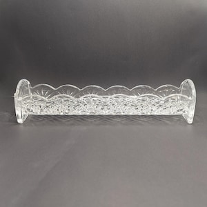 Op de afbeelding: Een heldere glazen schaal met een rechthoekige vorm en een decoratieve geschulpte rand. De schaal heeft een reli&euml;f bloemmotief en gebogen, verhoogde uiteinden. Geschikt voor serveren of presenteren. Afmetingen zijn ongeveer 25 cm lang.