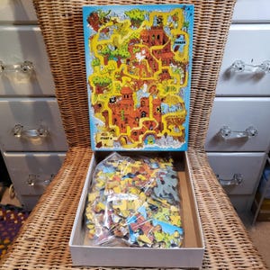 May include: A rectangular jigsaw puzzle box featuring a colorful illustrated maze with a start and finish point. The box lid displays a fantasy landscape with a castle, trees, and a volcano. Inside the open box, loose puzzle pieces are contained in a clear plastic bag.
