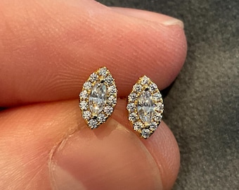 May include: A pair of gold-colored stud earrings. Each earring features a central marquise-cut clear stone, surrounded by a halo of smaller, round, clear stones. The earrings are held between fingers against a neutral background.