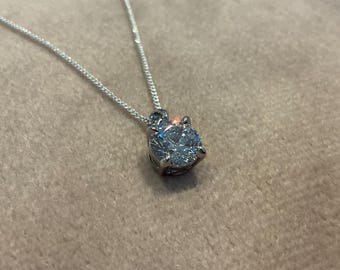 May include: A silver necklace with a large, round, clear gemstone pendant. The pendant is set in a four-prong setting and sparkles. The chain is delicate and silver. The necklace is displayed on a neutral-colored surface.