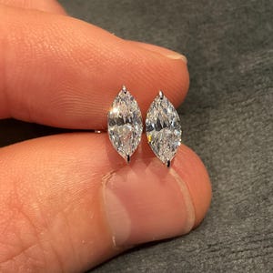 Puede incluir: Un par de pendientes de diamantes con corte marquesa. Los diamantes están engastados en una montura de plata u oro blanco. Los pendientes se sostienen entre dos dedos sobre un fondo gris.