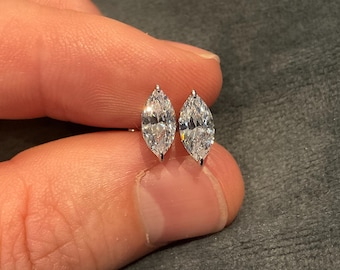 May include: A pair of marquise-cut diamond stud earrings. The diamonds are set in a silver or white gold setting. The earrings are held between two fingers against a grey background.